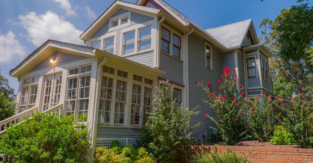 Two-story gray siding house with white trim, glass sunroom, brick wall, flowering shrubs.