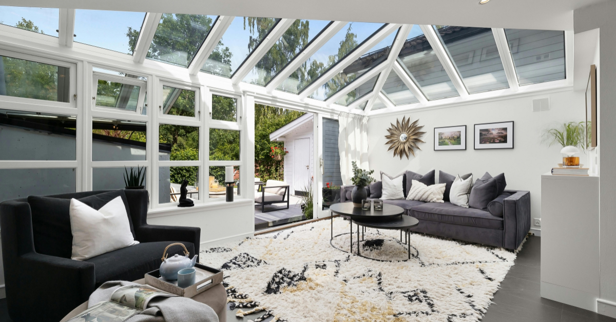 Sunlit glass-roof sunroom with gray sofa, black armchair, and cream shag rug.
