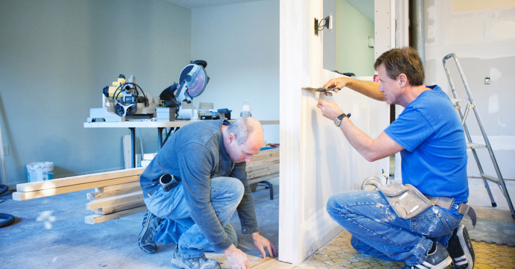 Two contractors remodeling; one trims wood planks, the other tapes a drywall seam.