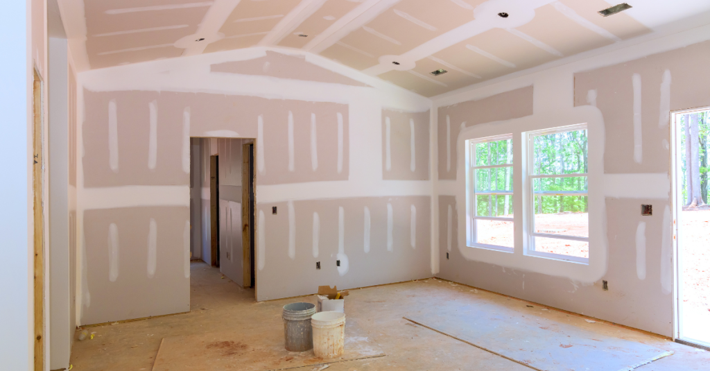 Unfinished drywall room with taped joints, two windows, and buckets on the floor.