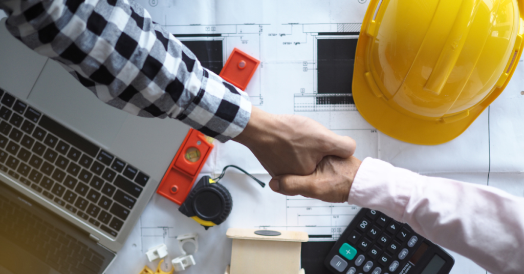 Two people shake hands over blueprints with a yellow hard hat, level, calculator, laptop